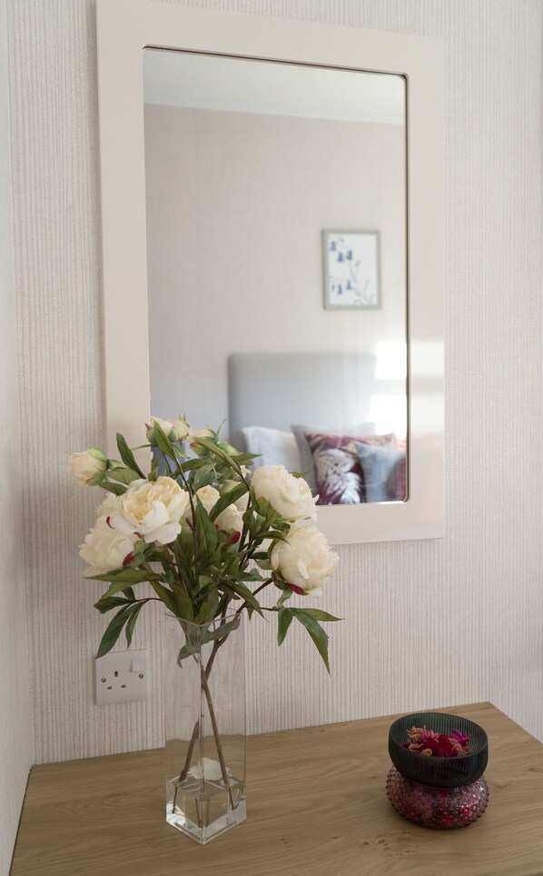 A clear glass vase with white flowers on a wooden table, a mirror on the wall, and a small decorative bowl with red flowers. - Hollicarrs - Bluebell Lodge (York)