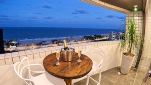 Ein Balkon mit einem Holztisch, zwei weißen Stühlen, einem Topfpflanzen und Blick auf den Ozean und den Strand im Abenddämmerung.