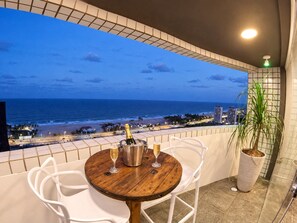 A balcony with a wooden table, two white chairs, a potted plant, and a view of the ocean and beach at dusk. - Cobertura vista mar com deck privativo (Fortaleza)