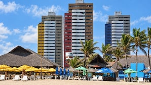 Ein Strand mit strohgedeckten Hütten und bunten Sonnenschirmen, feiner Sand und hohe Gebäude im Hintergrund.