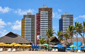 A beach with thatched-roof huts and colorful umbrellas, a sandy beach, and tall buildings in the background. - Cobertura vista mar com deck privativo (Fortaleza)