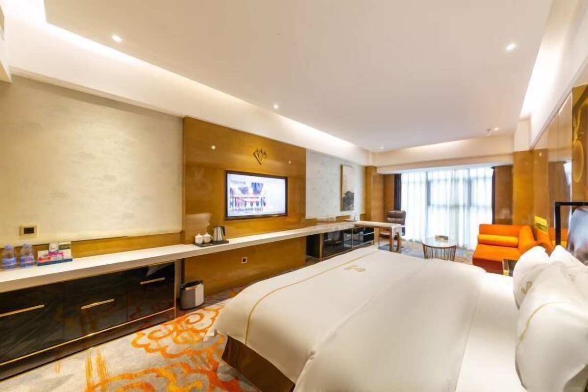 A hotel room with a large bed, a desk with a flat-screen TV, a small seating area, and a view of the city.