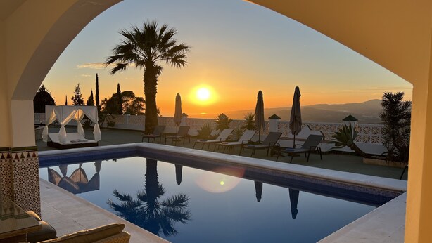 Un espace piscine doté de chaises longues et d’un lit à baldaquin, offrant une vue sur un coucher de soleil avec des montagnes en arrière-plan.