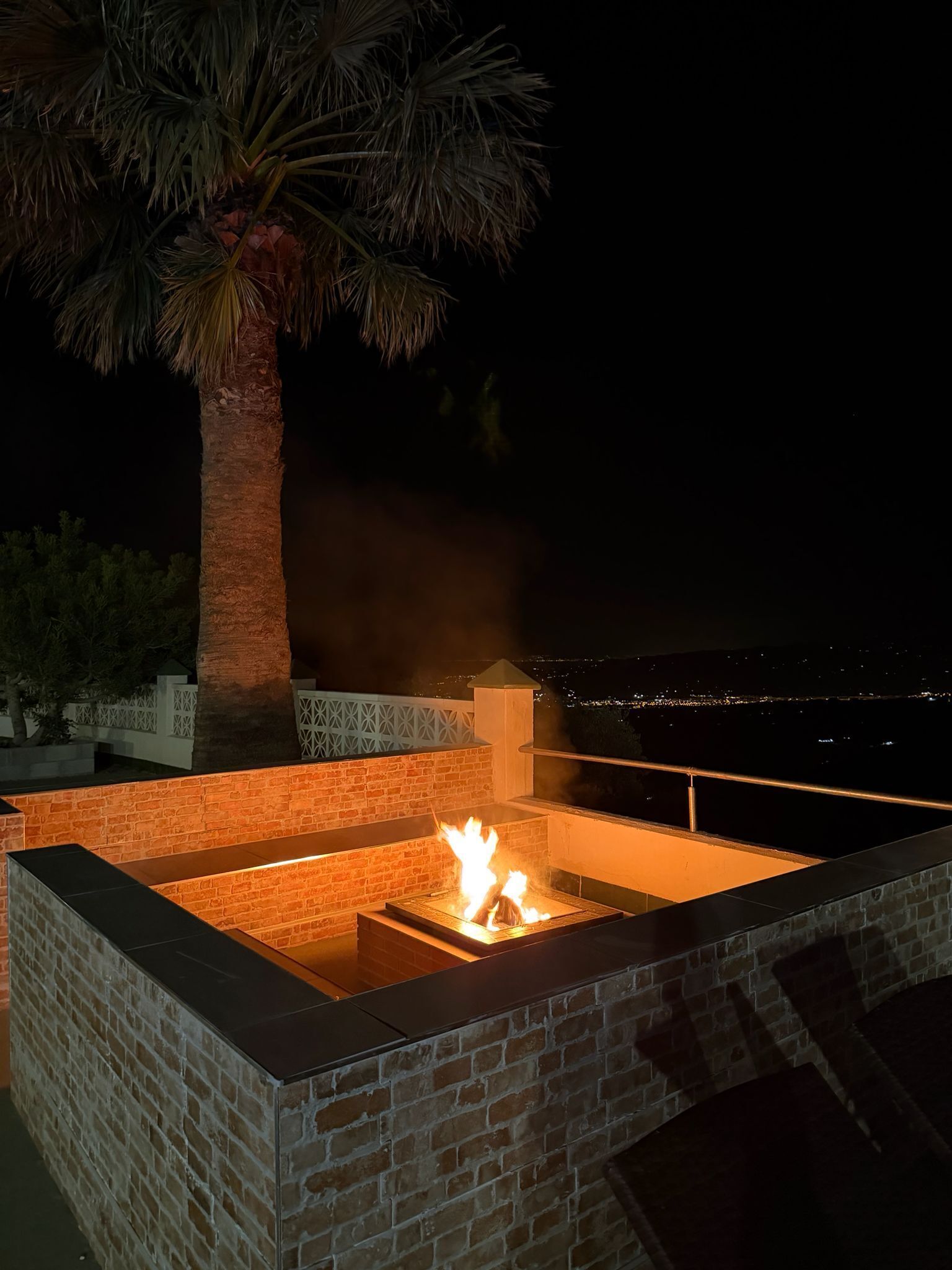 Una fogata al aire libre con un revestimiento de ladrillo, una palmera y vistas de un paisaje urbano nocturno.