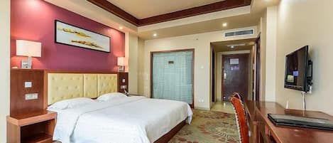 Quarto de hotel com uma cama grande, uma escrivaninha, uma cadeira, uma televisão e uma porta que dá para outro cômodo.
