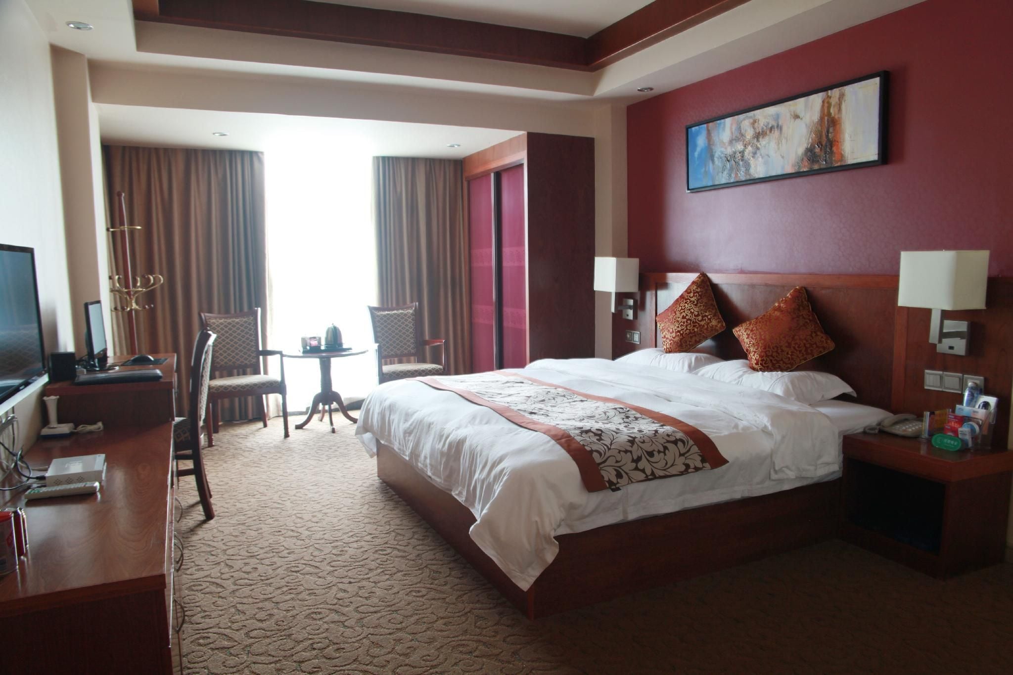 A hotel room with a large bed, a desk with a computer, a chair, a small table with a lamp, and a painting on the wall.
