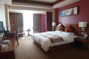 Habitación de hotel con una cama grande, un escritorio con una computadora, una silla, una mesita con una lámpara y un cuadro en la pared.