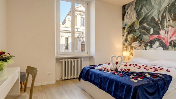 A hotel room with a bed, a chair, a table, a window with a view of buildings, and a wall with a floral painting. - Desenzanoloft Apa Dom  (Desenzano del Garda)