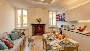 A modern living room with a sofa, a fireplace, a dining area with a table set for breakfast, and a kitchen with a TV displaying the BBC News. - Desenzanoloft Apa Dom  (Desenzano del Garda)