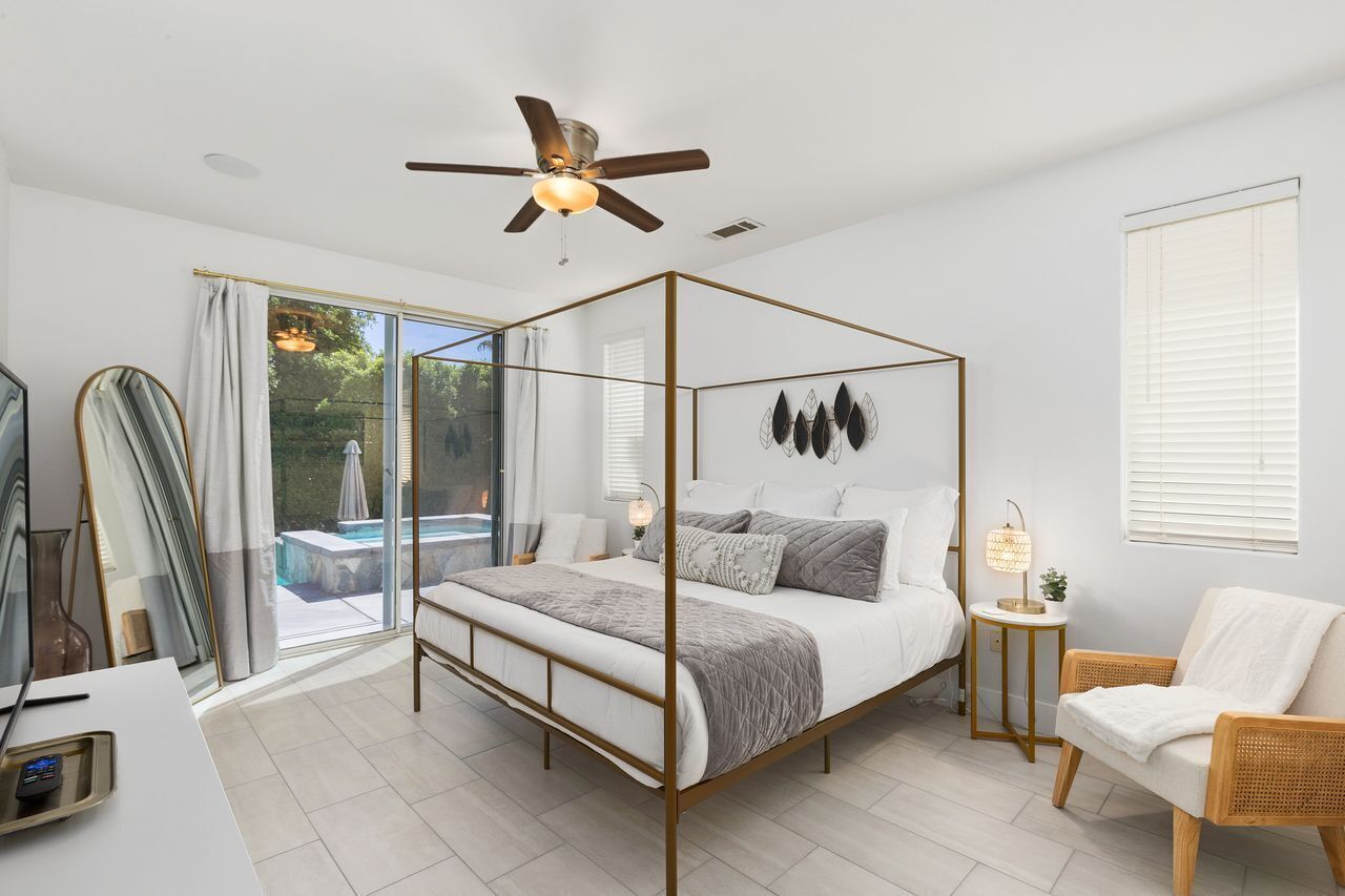 A modern bedroom with a four-poster bed, a ceiling fan, a bedside table with a lamp, and a view of a swimming pool through glass doors.