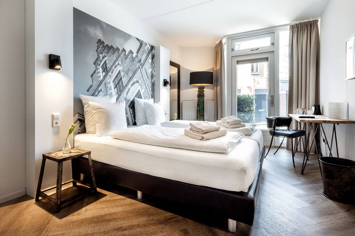 A modern hotel room with a large bed, a small side table, a desk with a chair, and a view of the city through the window.