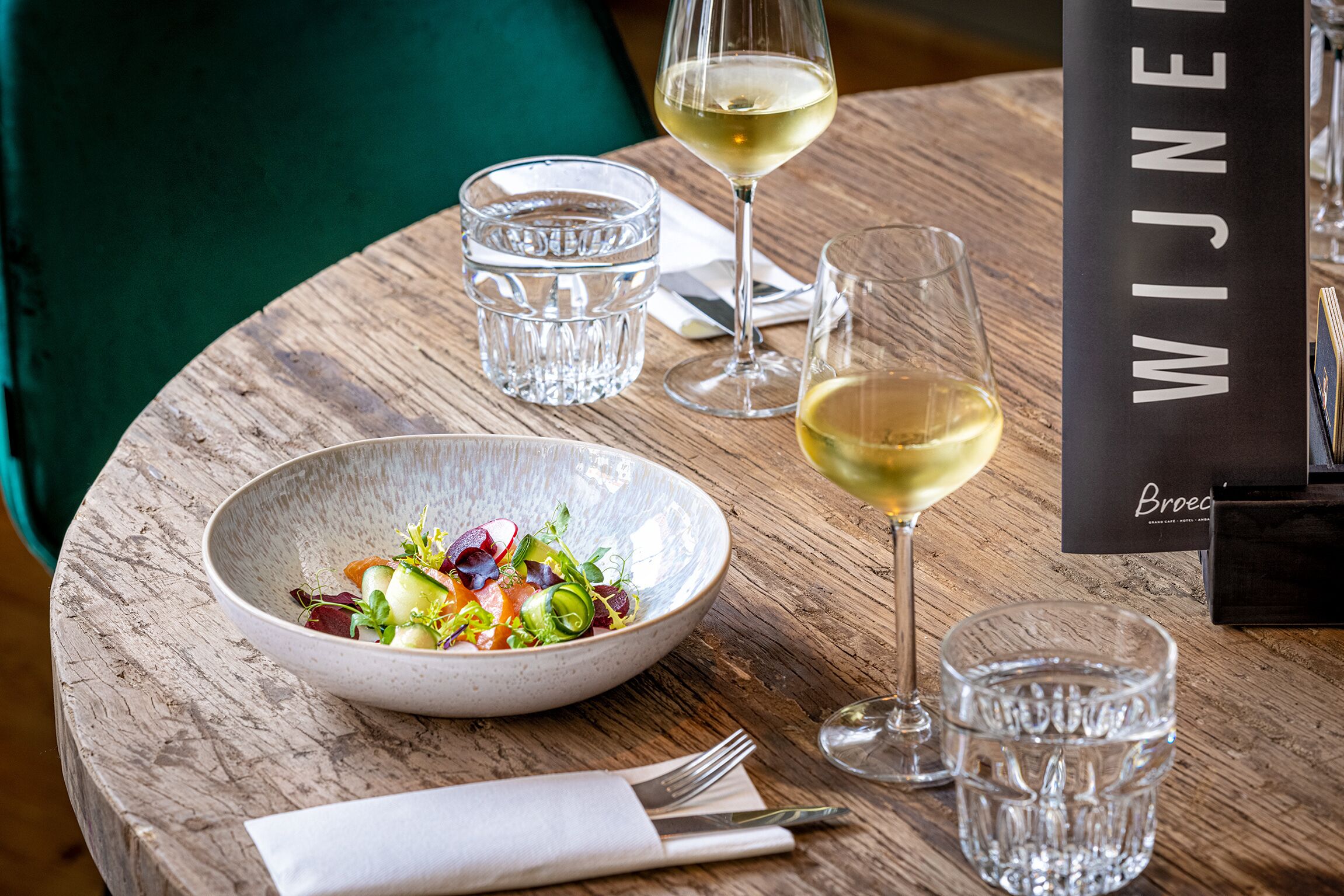Et træbord med en salatskål, to glas hvidvin og en menu med teksten 'WIJNEN BROEO'.