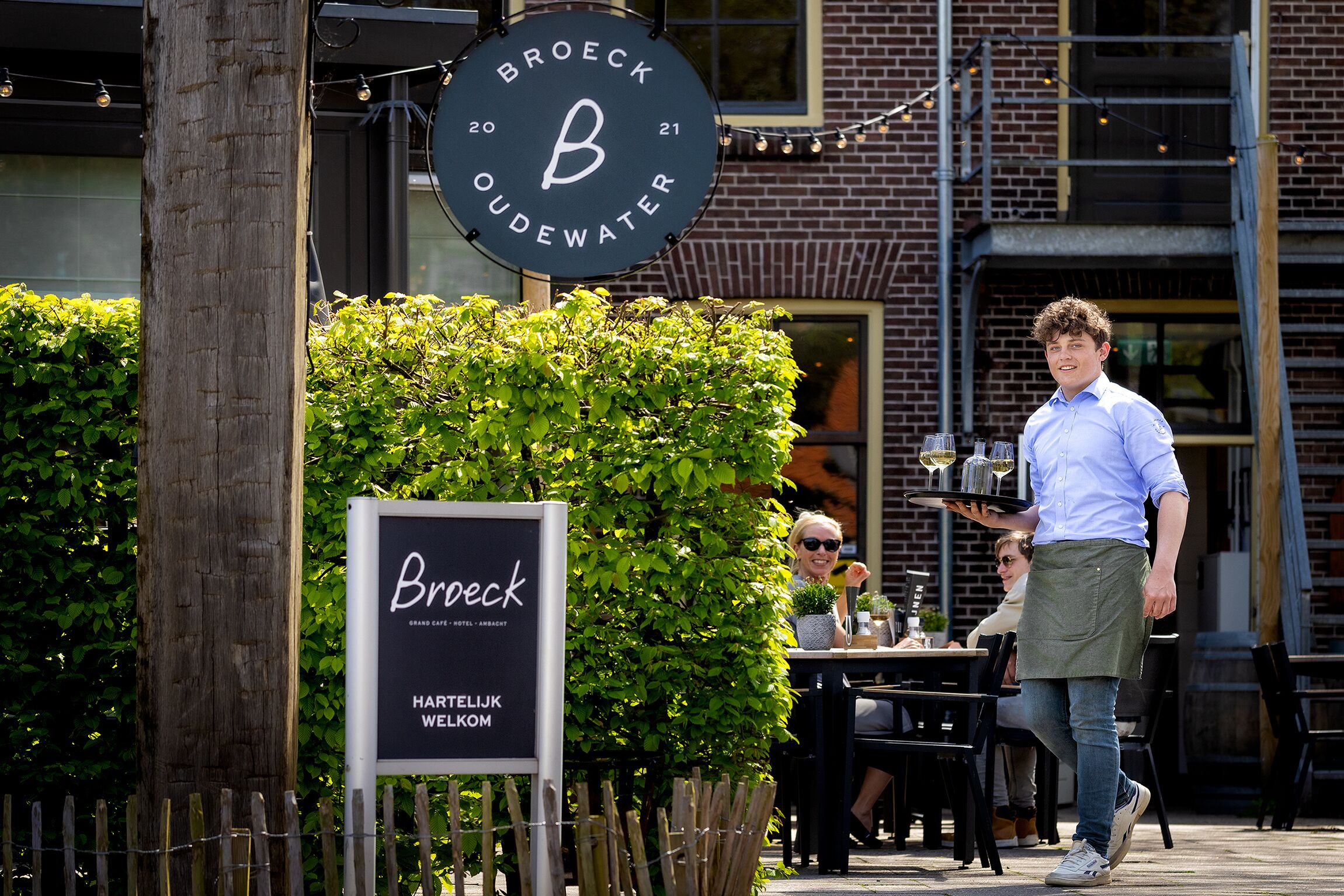 A sign for "Broeck Oudewater" with a large 'B' logo, a wooden post, and a hedge in the foreground.