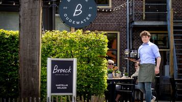 A sign for "Broeck Oudewater" with a large 'B' logo, a wooden post, and a hedge in the foreground.