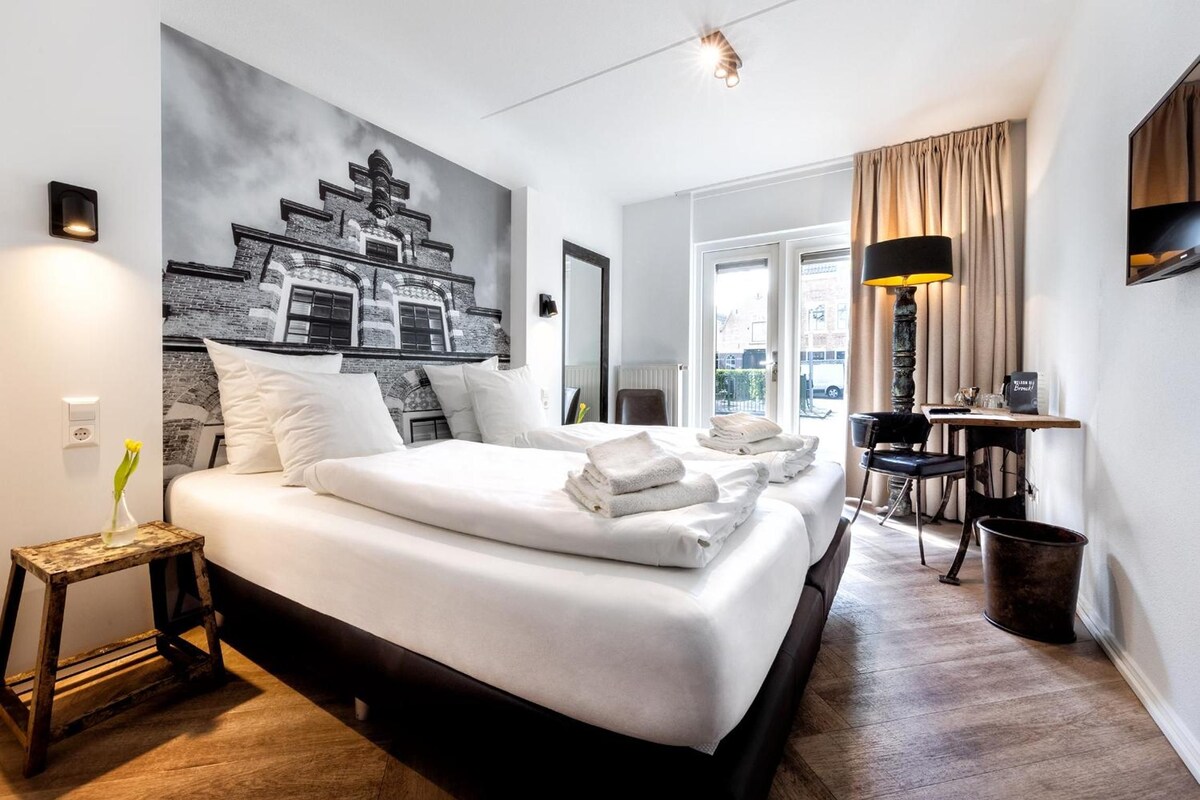 A hotel room with a large bed, a desk, a chair, a lamp, and a view of the outside.