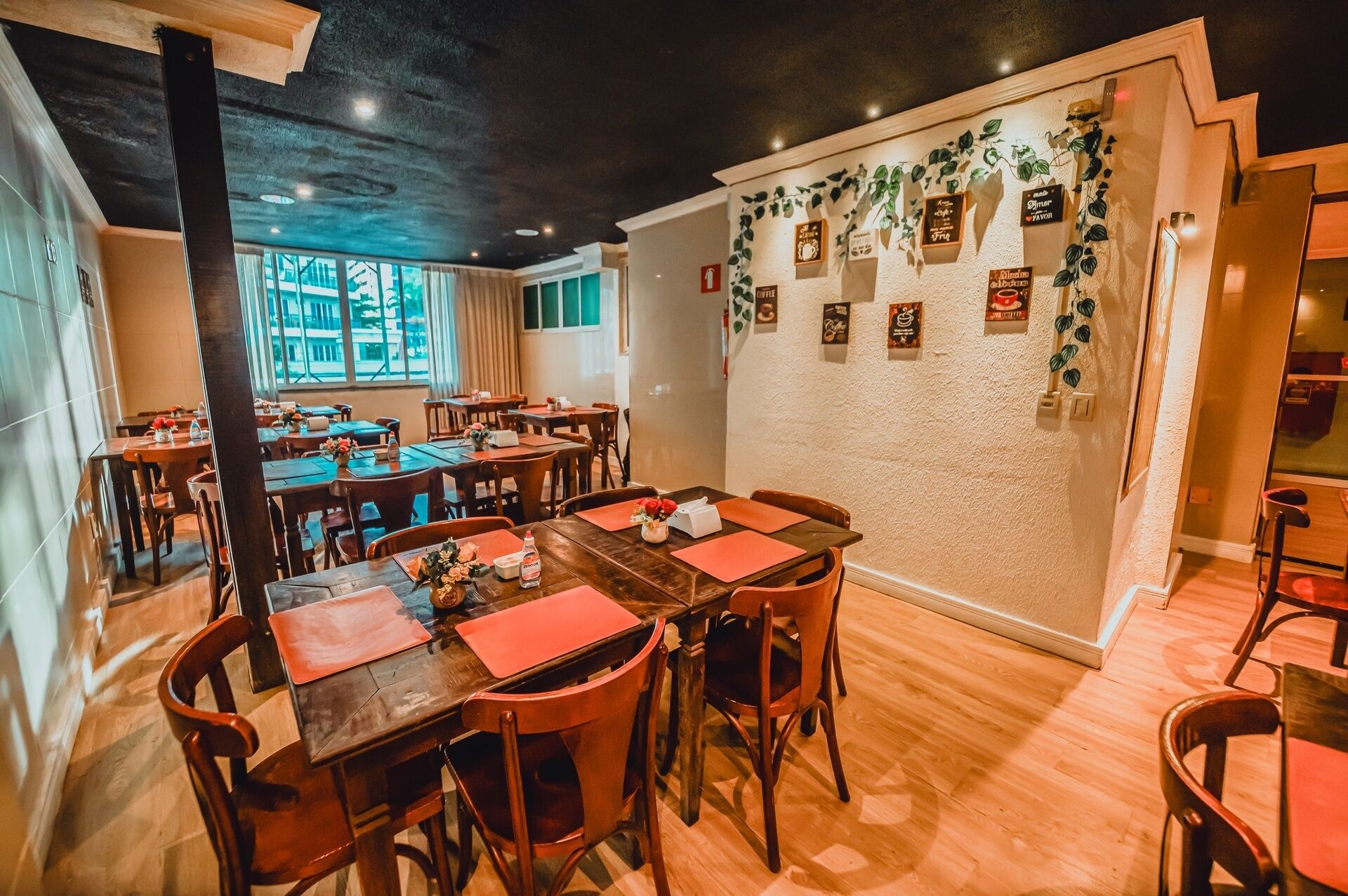 Un restaurant avec des tables et des chaises en bois, un mur blanc à la texture particulière orné de tableaux encadrés, et une vue dégagée grâce à de grandes fenêtres.