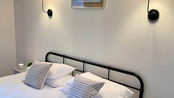 Una cama bien hecha con sábanas blancas y almohadas a rayas, un cuadro en la pared y dos lámparas fijadas a la pared.