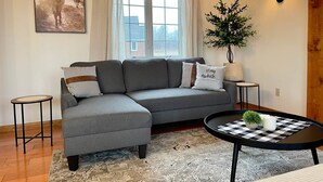 Een moderne woonkamer met een grijze hoekbank, een ronde salontafel, een kamerkleed met dessin, een ingelijst kunstwerk aan de muur en een potplant.