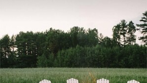 Een cirkelvormige opstelling van witte Adirondack-stoelen rond een vuurkuil met stenen en houtblokken, gelegen op een grindpad in een grasveld met bomen op de achtergrond.