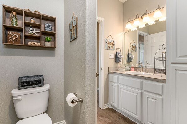 A bathroom with a toilet, a shelf with decorative items, a mirror with lights, and a sink with a vanity. - 4-bedroom family home walking distance to the beach. 27 miles of golf cart fun. (Bolivar Peninsula)