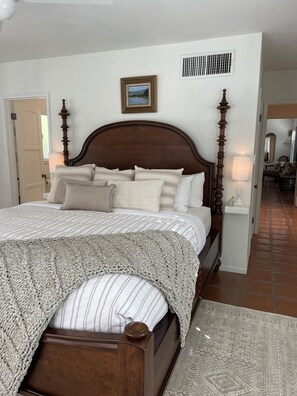 A neatly made bed with a wooden headboard, a patterned throw blanket, and a view of a hallway leading to another room.