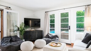 A modern living room with a flat-screen TV, a dark wooden cabinet, a white sofa, and a large window with a view of a swimming pool. - Historical 1937 Movie Colony Spanish Hacienda (Palm Springs)