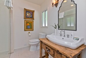 A bathroom with a white sink, wooden vanity, two framed paintings, a toilet, and a window.