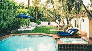A swimming pool with lounge chairs and umbrellas, surrounded by greenery and a white building.