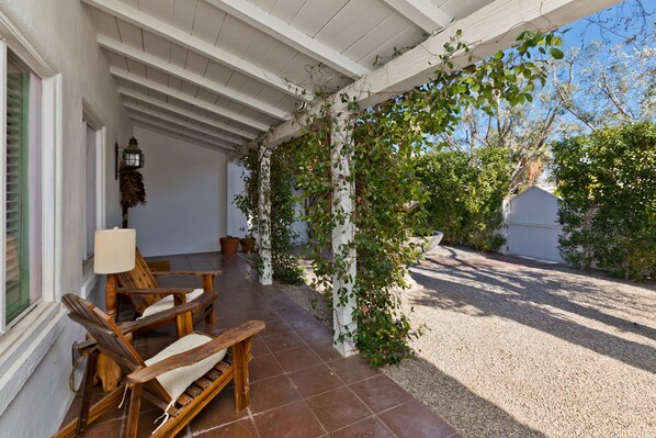 A covered patio with wooden chairs and a table, a white wall, and a garden area with gravel and greenery.