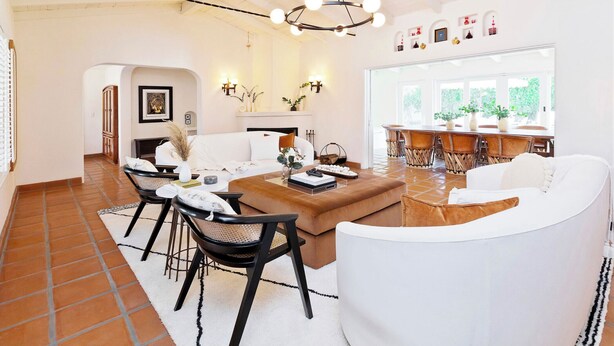 A spacious living area with a large white sofa, a brown ottoman, and a dining table with chairs. There is a fireplace with a mantel and wall-mounted sconces. The room has a high ceiling with exposed beams and a large window providing natural light. - Historical 1937 Movie Colony Spanish Hacienda (Palm Springs)
