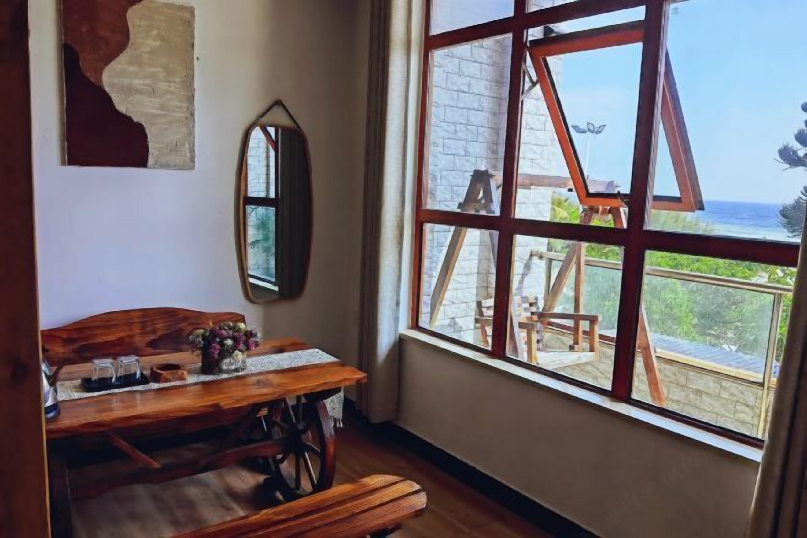 A room with a wooden table, a bench, a mirror, and a large window with a view of the ocean.