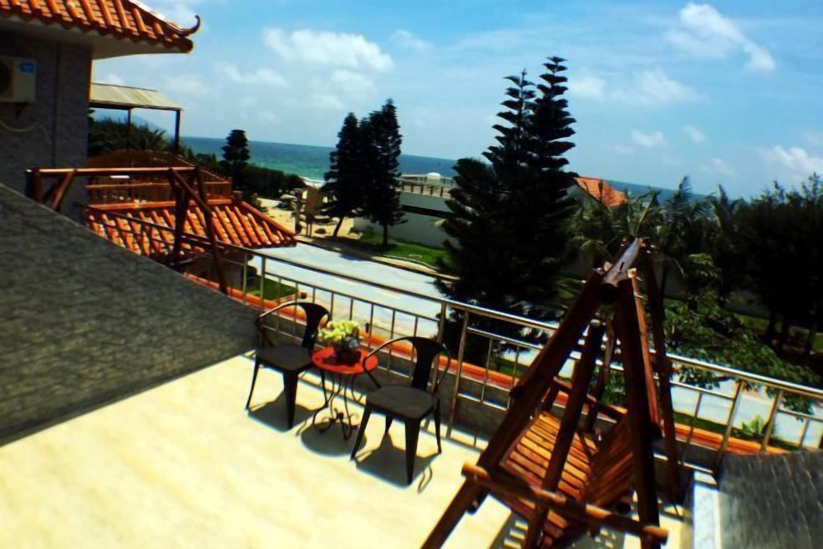 A balcony with a view of the ocean, surrounded by tropical trees and a building with a tiled roof.