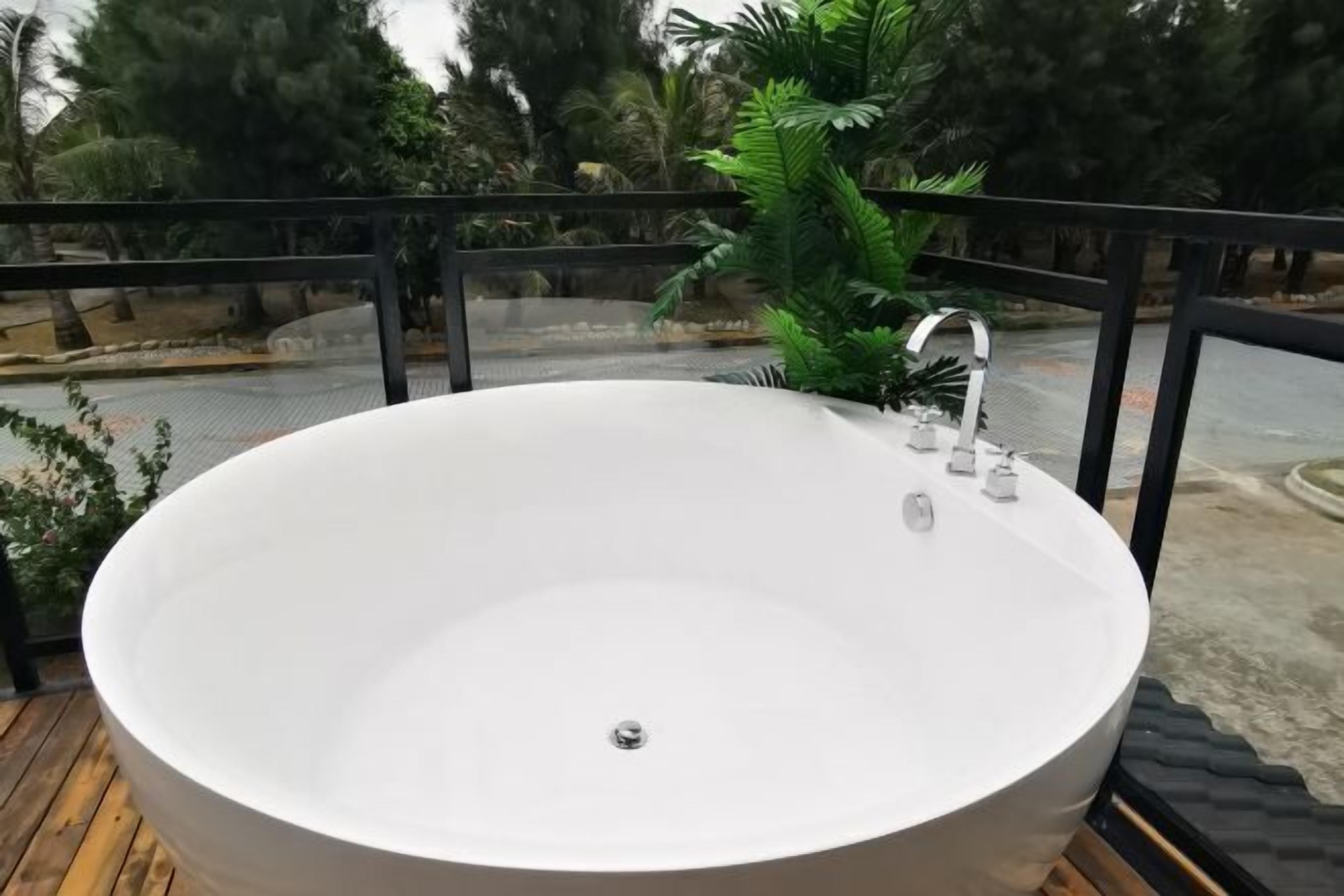 A large, round, white bathtub on a wooden deck with a black railing, surrounded by greenery.