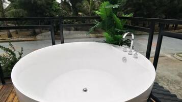 A large, round, white bathtub on a wooden deck with a black railing, surrounded by greenery.