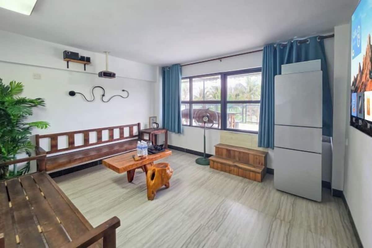Een kamer met een houten bankje, een tafel, een koelkast, een raam met gordijnen en een plant.