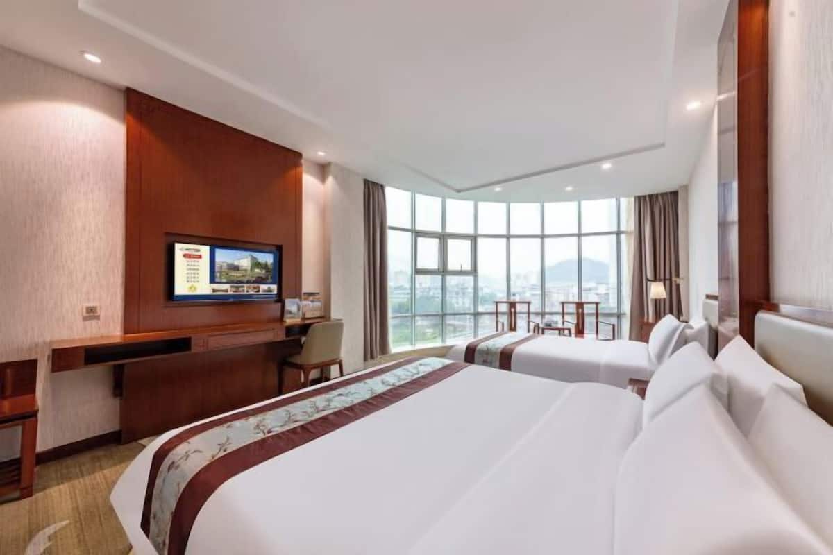 A hotel room with two beds, a desk, a TV, and a large window with a city view.
