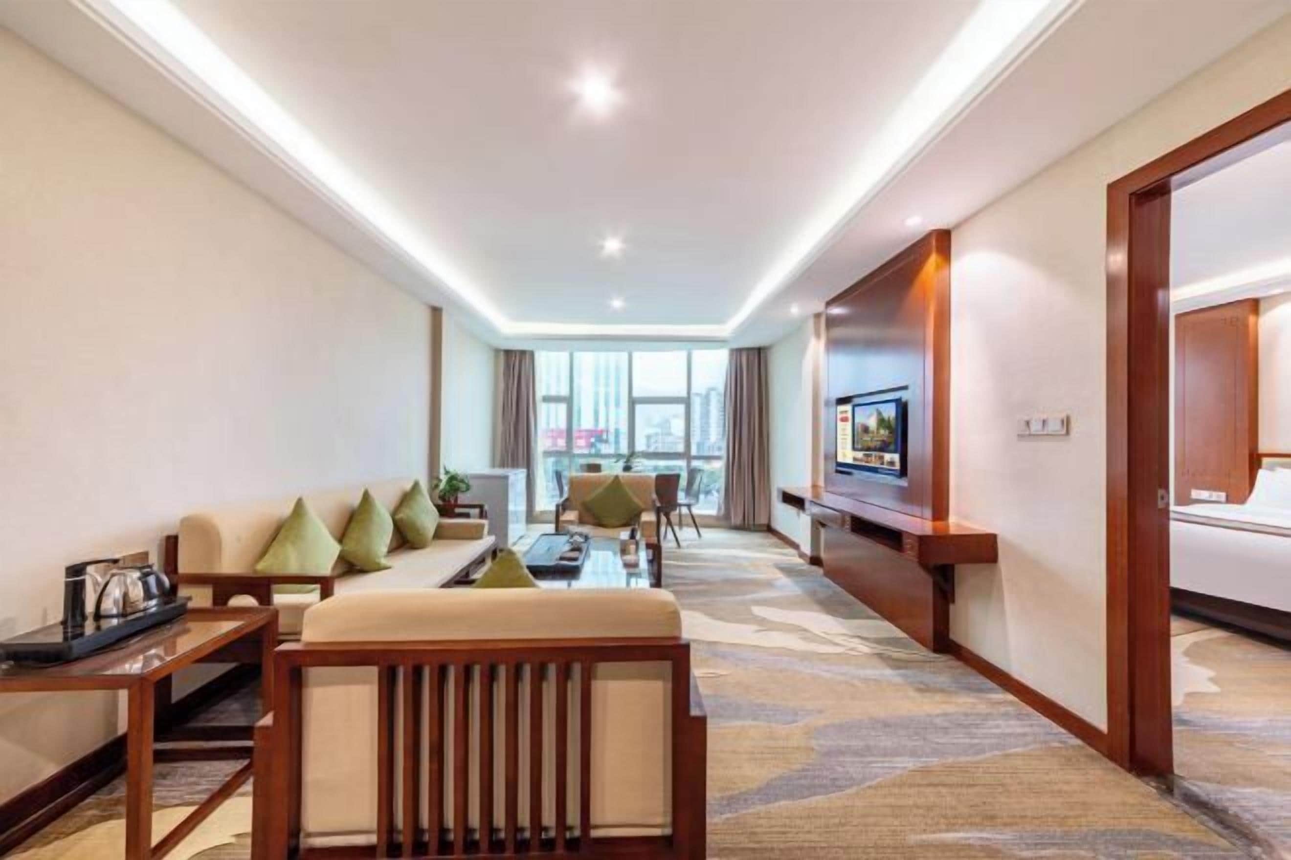 A hotel room with a sofa, armchairs, a coffee table, a TV, and a large window.