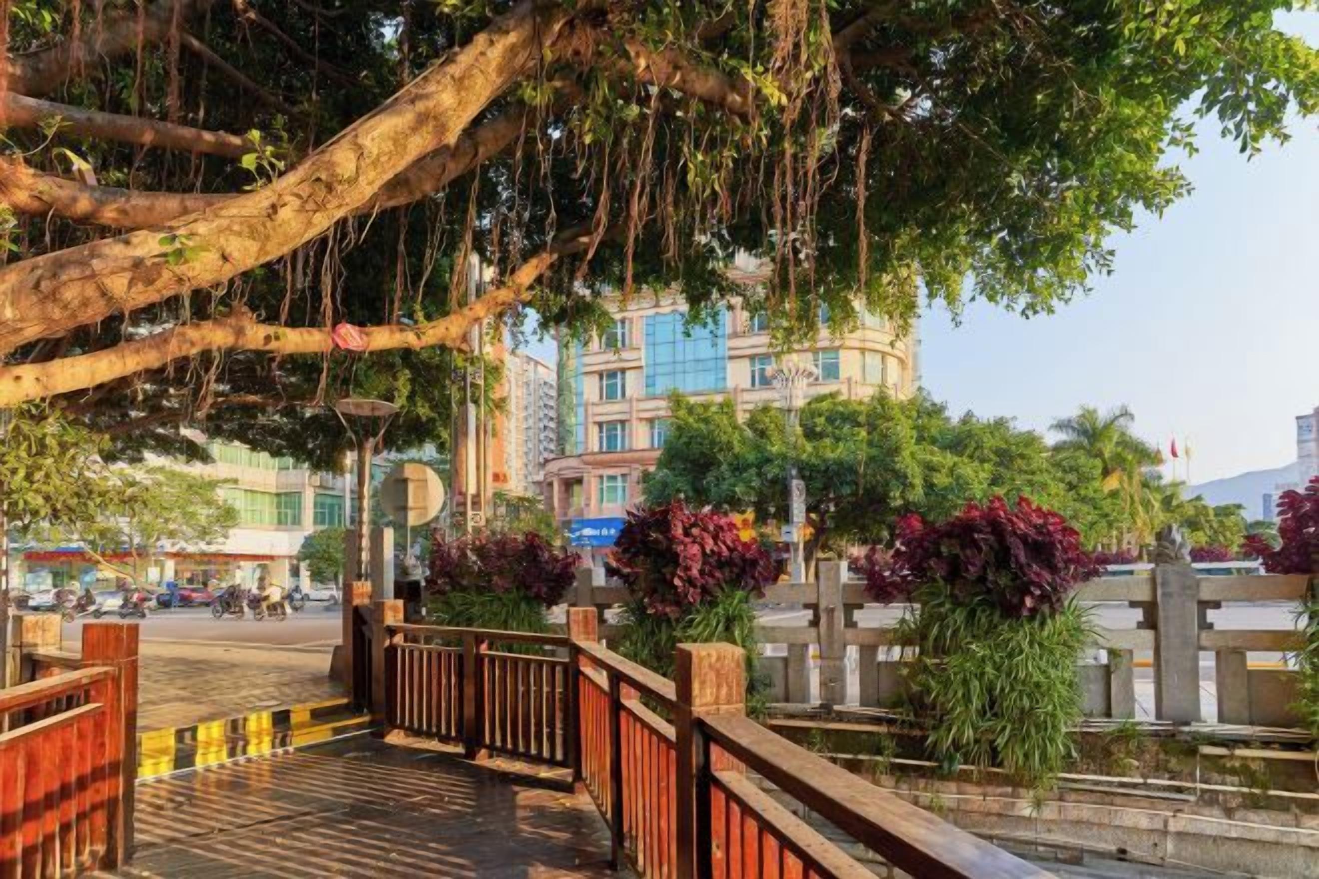 A pedestrian bridge with a wooden railing, surrounded by greenery and buildings in the background.
