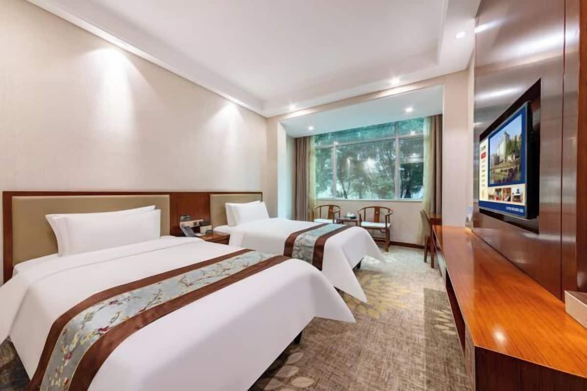 A hotel room with two beds, a desk, a TV, and a large window with a view of trees.