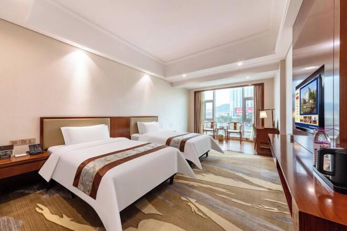 A hotel room with two beds, a desk, a chair, a TV, and a large window.