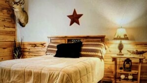 A rustic bedroom with a wooden bed, a mounted deer head, a star decoration, and a nightstand with a lamp. - Cozy Cabin with Beautiful Views. Minutes from Downtown Custer (Custer)