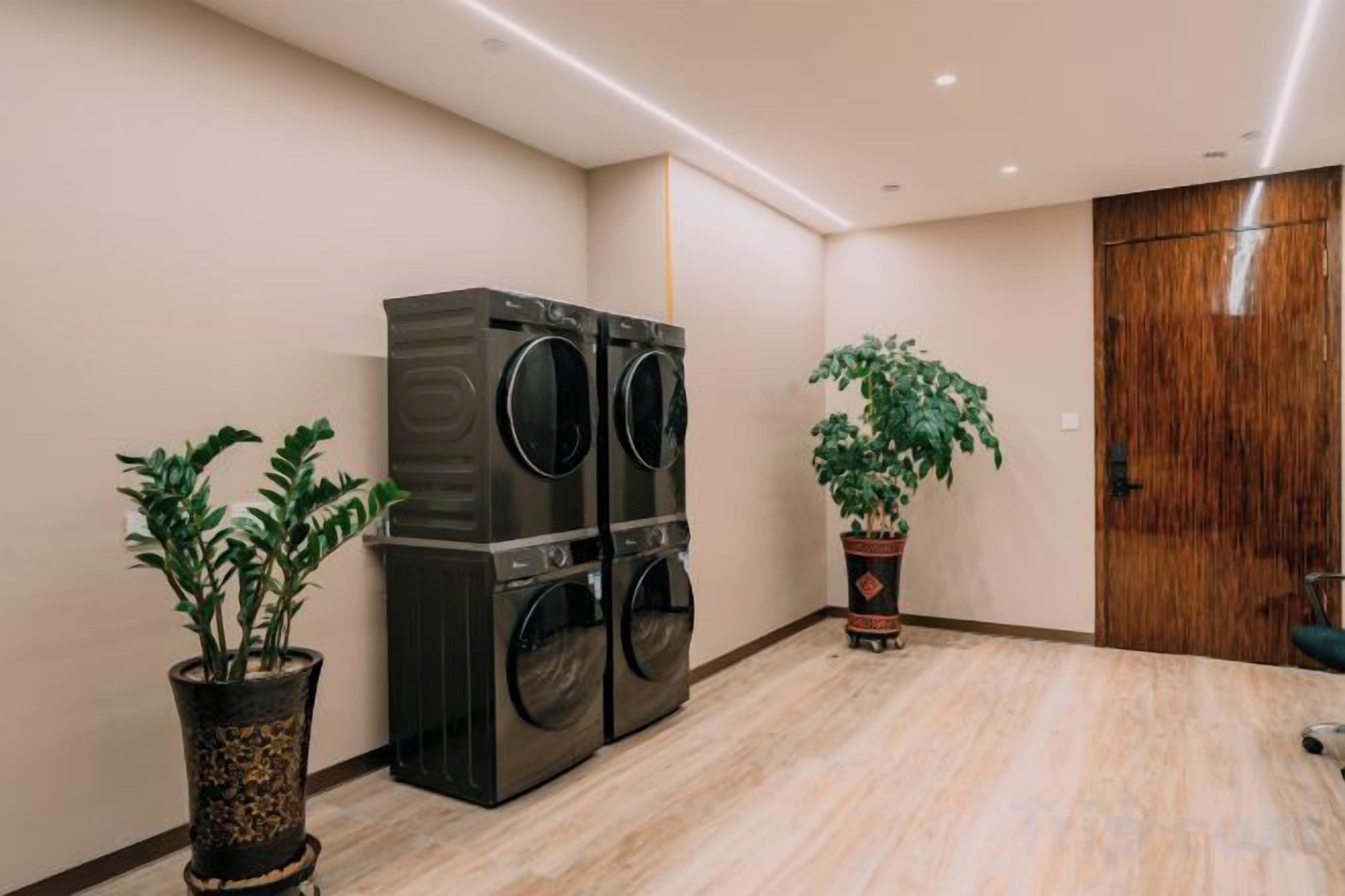 Uma lavanderia com uma máquina de lavar e uma secadora grandes, duas plantas em vasos e uma porta de madeira.