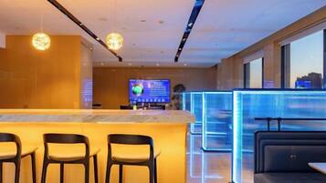 A modern hotel lobby with a reception desk, bar area, seating, and a large screen displaying information.