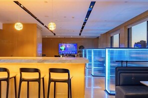 Una hall d'albergo moderna con reception, area bar, posti a sedere e un grande schermo che mostra informazioni.