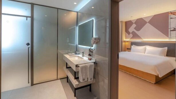 A modern hotel room with a glass partition, a double sink vanity, a large bed, and a wall-mounted mirror. - Shangshi Hotel (Fuyun)