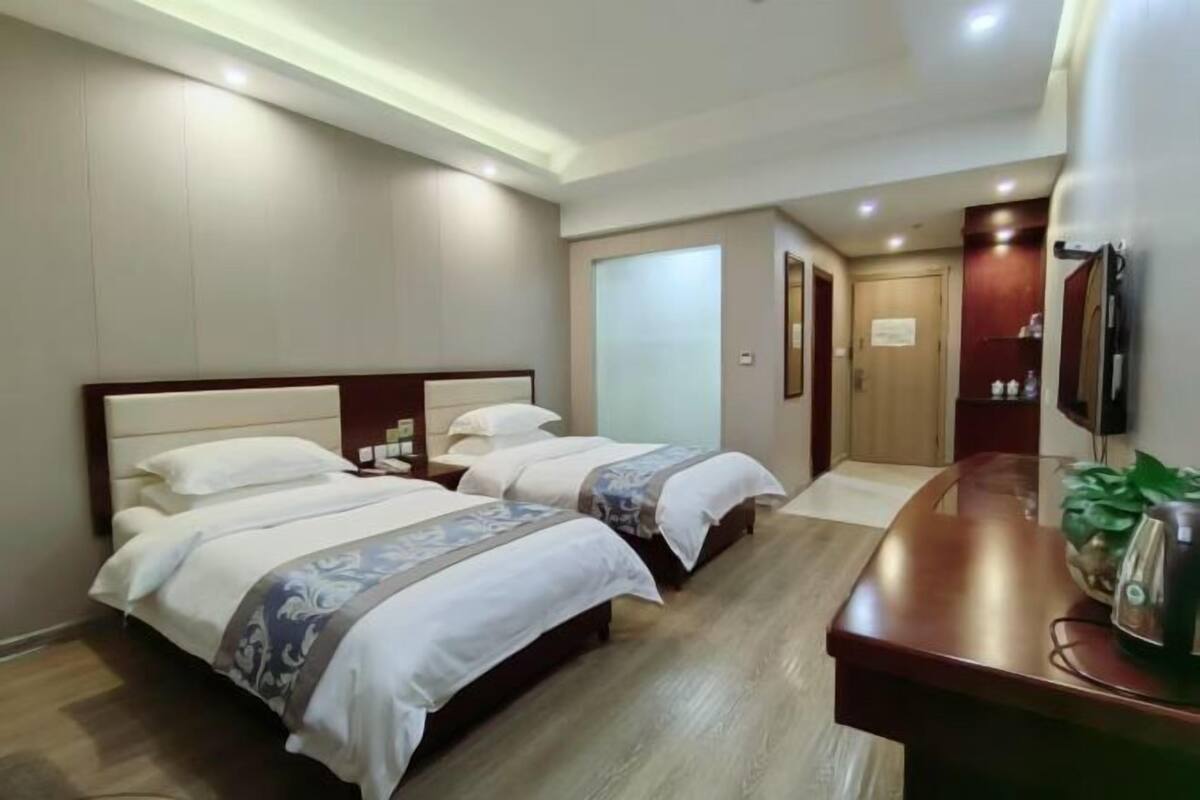 A hotel room with two beds, a wooden floor, a desk with a plant, and a TV on the wall.