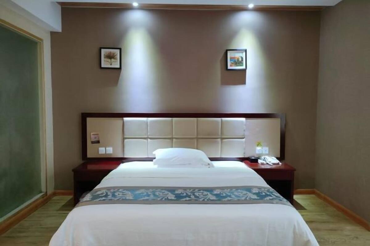 A hotel room with a large bed, a headboard with a light panel, a bedside table with a phone, and two framed pictures on the wall.