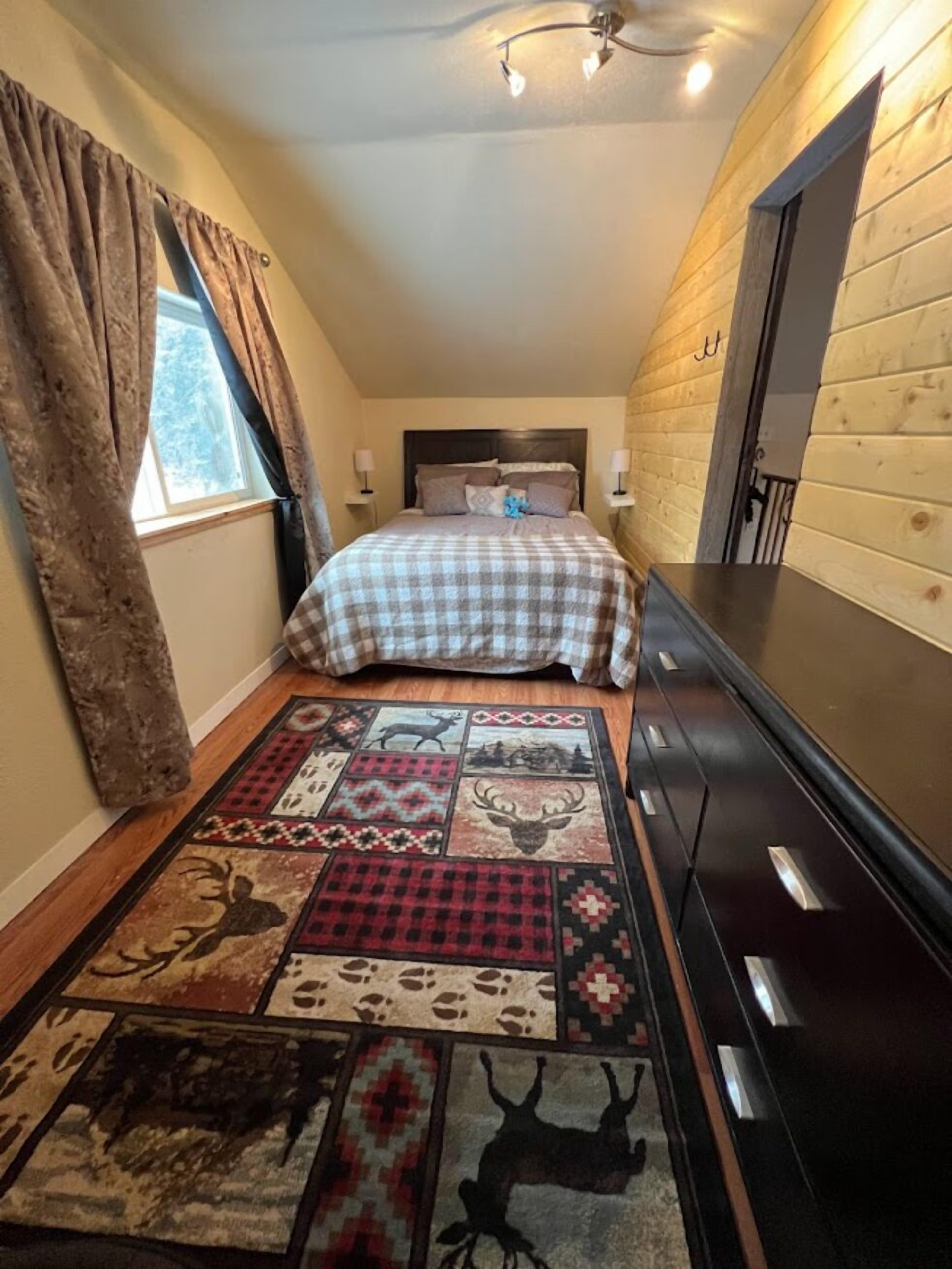 A bedroom with a checkered bedspread, a patterned rug, a window with curtains, a headboard, bedside lamps, and a dresser.