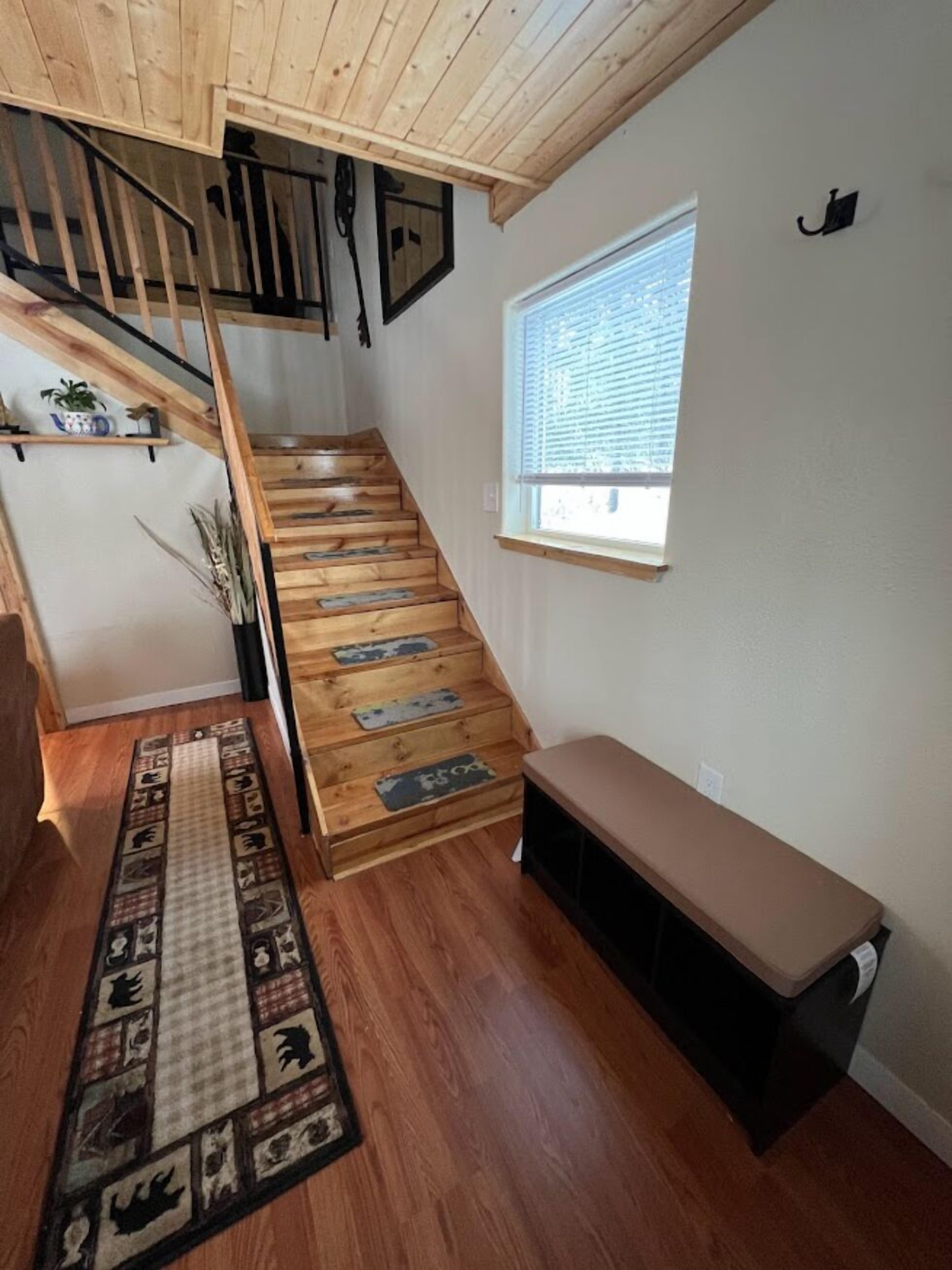 A wooden staircase with a bench at the bottom, a window with blinds, and a patterned rug on the floor.