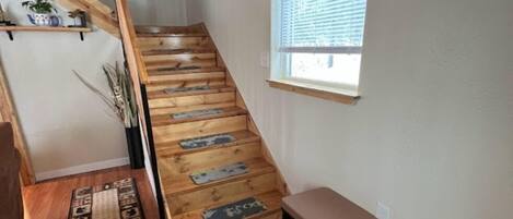 A wooden staircase with a bench at the bottom, a window with blinds, and a patterned rug on the floor.